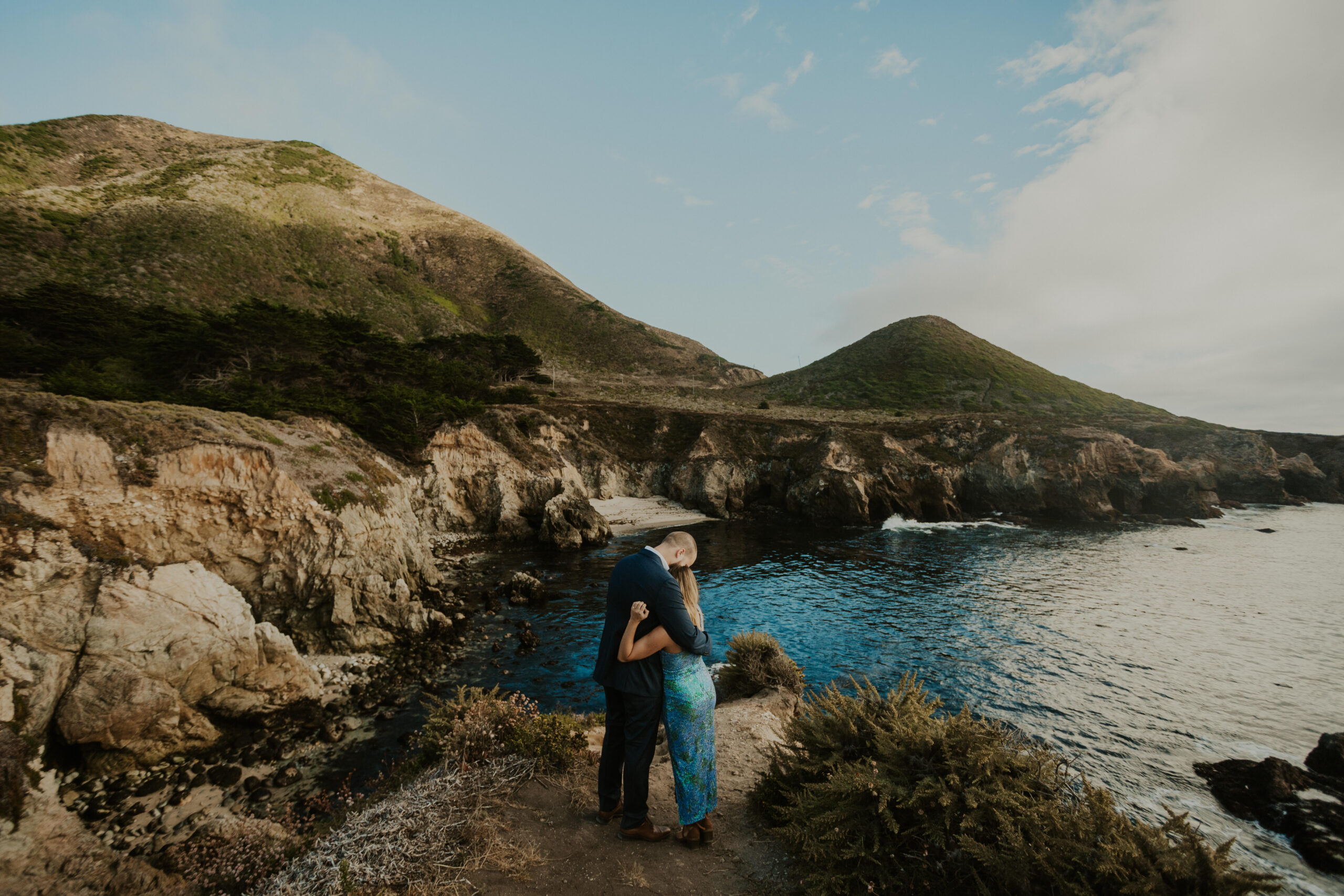 A couple dressed fancy apparel hugs overlooking a beautiful coastal landscape of rocks and hills and water at their Big Sur proposal.