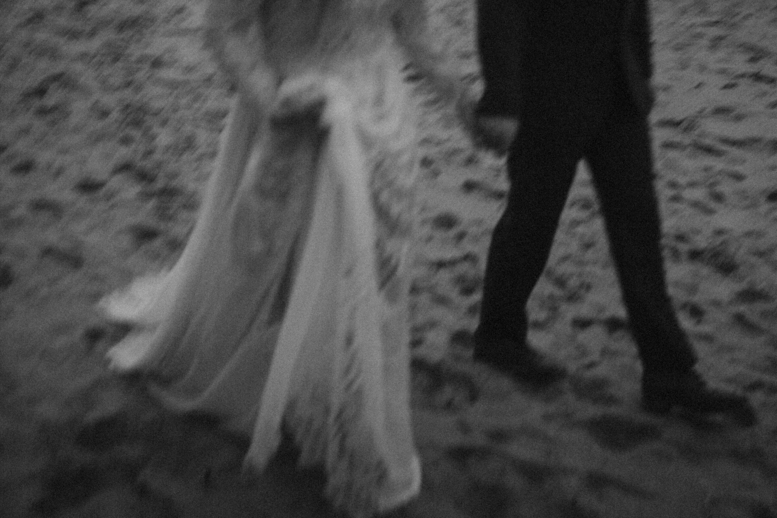 a bride holds up her dress as she and her husband walk along the sand. black and white image, slightly blurred. they just had an intentional elopement