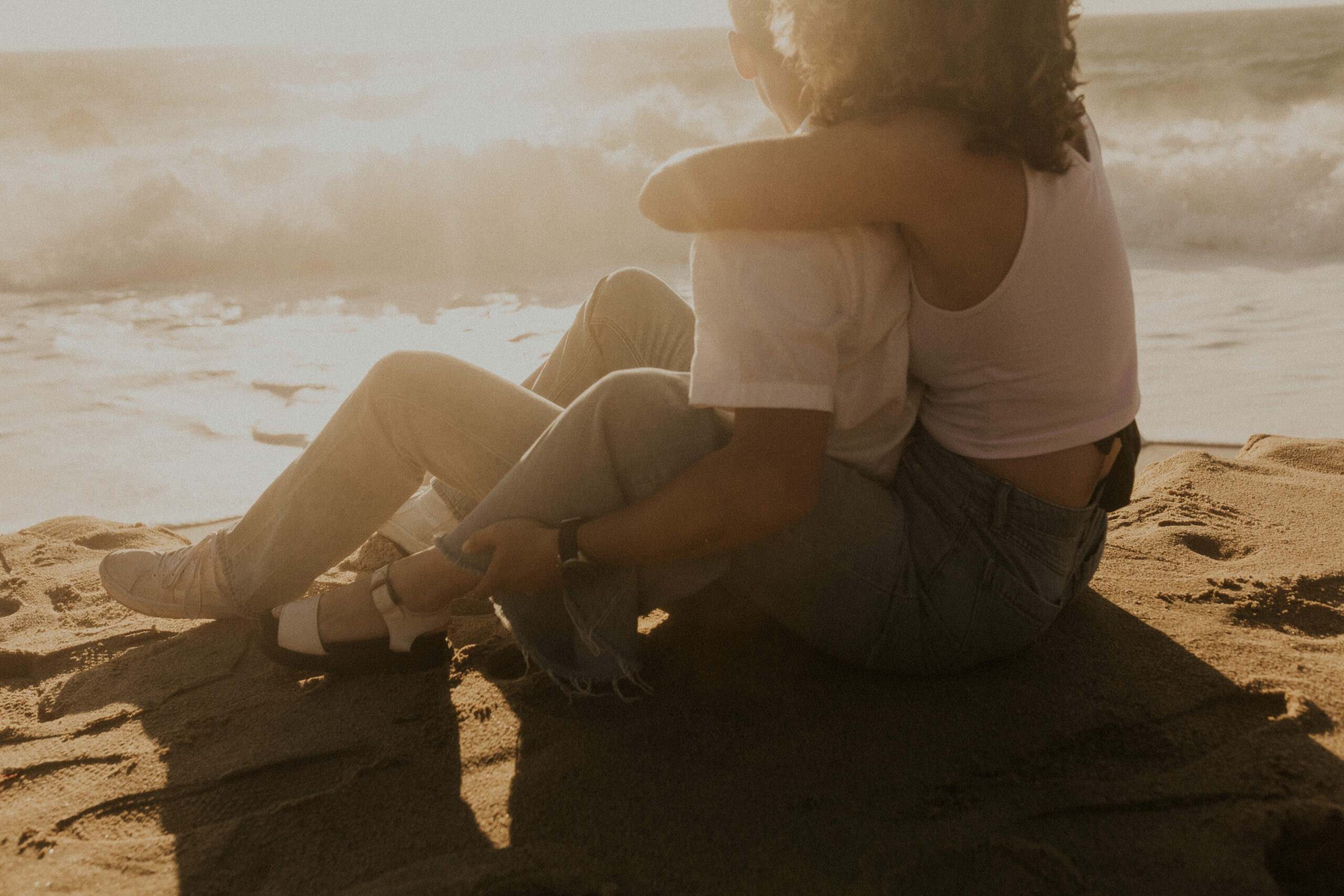 a couple sits on the beach, looking at the sunset over the ocean. they hug each other. this image is from a photo shoot in big sur, california that they booked with local photographer, paper airplanes photography