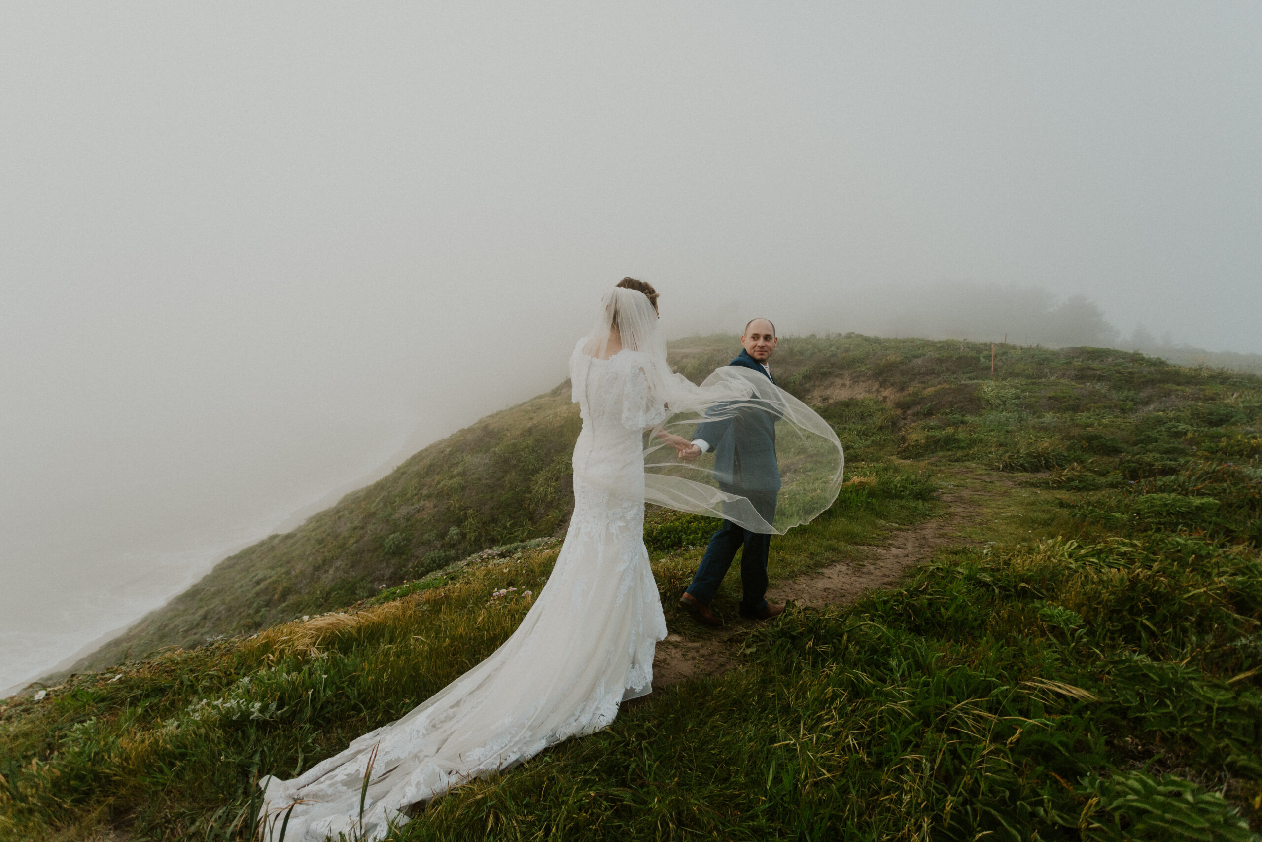 groom leads bride along a path on a green cliff at their intimate elopement